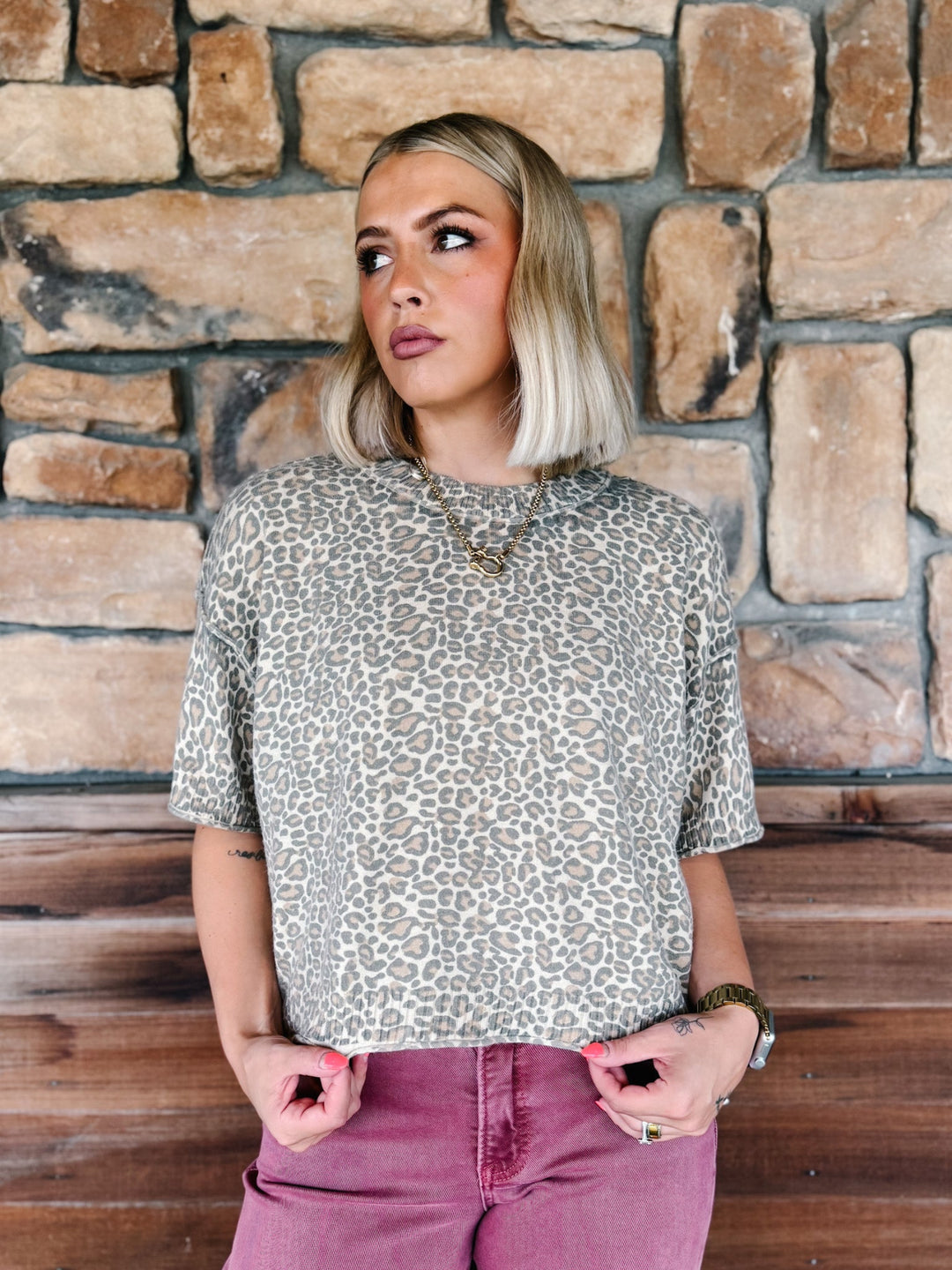 A woman with blonde bob-cut hair wearing a beige and brown leopard print short-sleeve top and dusty rose pants, standing in front of a rustic stone wall.