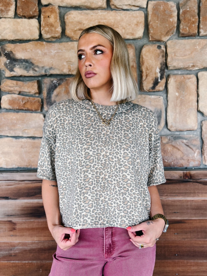 A woman with blonde bob-cut hair wearing a beige and brown leopard print short-sleeve top and dusty rose pants, standing in front of a rustic stone wall.