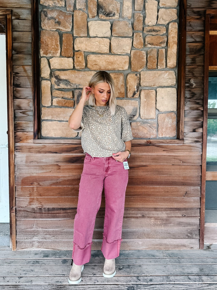 A woman stands on a rustic wooden porch in front of a stone wall, wearing a beige patterned short-sleeve top, dusty rose wide-leg jeans, and beige boots.