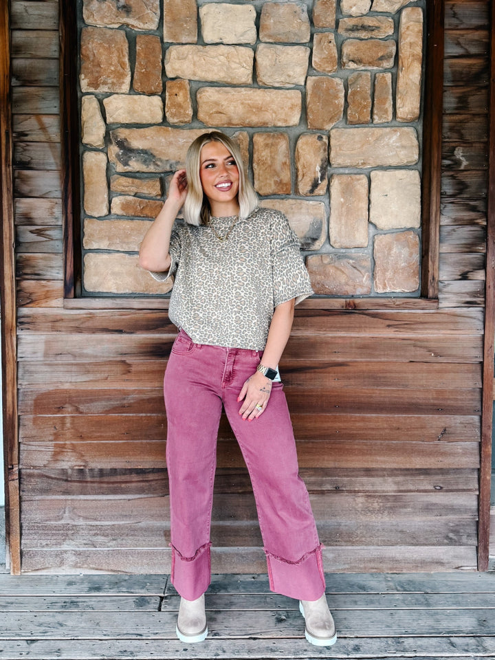A woman stands on a wooden porch in front of a stone wall, wearing a beige patterned short-sleeve top and dusty rose wide-leg cropped pants with rolled cuffs.