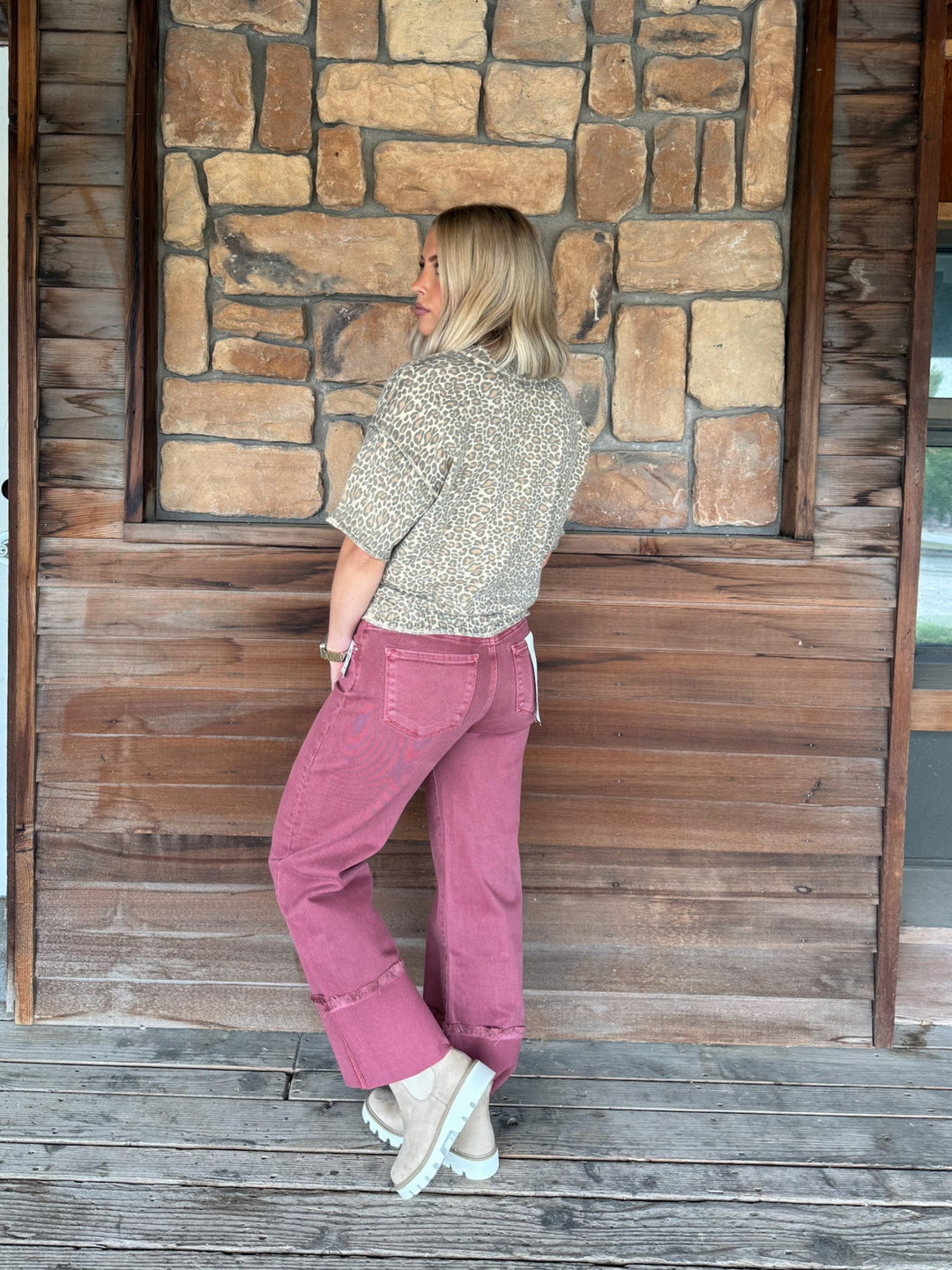 A woman stands on a wooden porch in front of a stone wall, wearing a beige patterned short-sleeve top and dusty rose wide-leg jeans with white sneakers.
