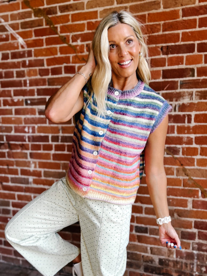 A woman posing in front of a red brick wall, wearing a colorful striped knit sleeveless vest with white buttons and cream-colored polka dot pants.