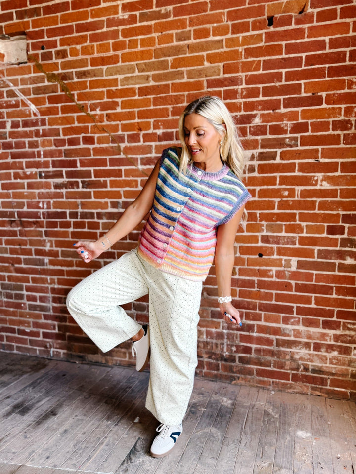 A woman posing in front of a red brick wall, wearing a colorful striped sleeveless top, cream-colored wide-leg pants, and white sneakers.