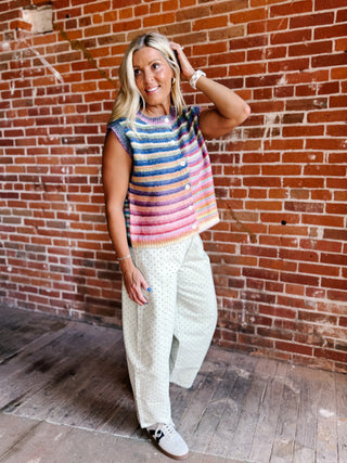 A woman posing in front of a red brick wall, wearing a colorful striped sleeveless knit top, white wide-leg patterned pants, and white sneakers.