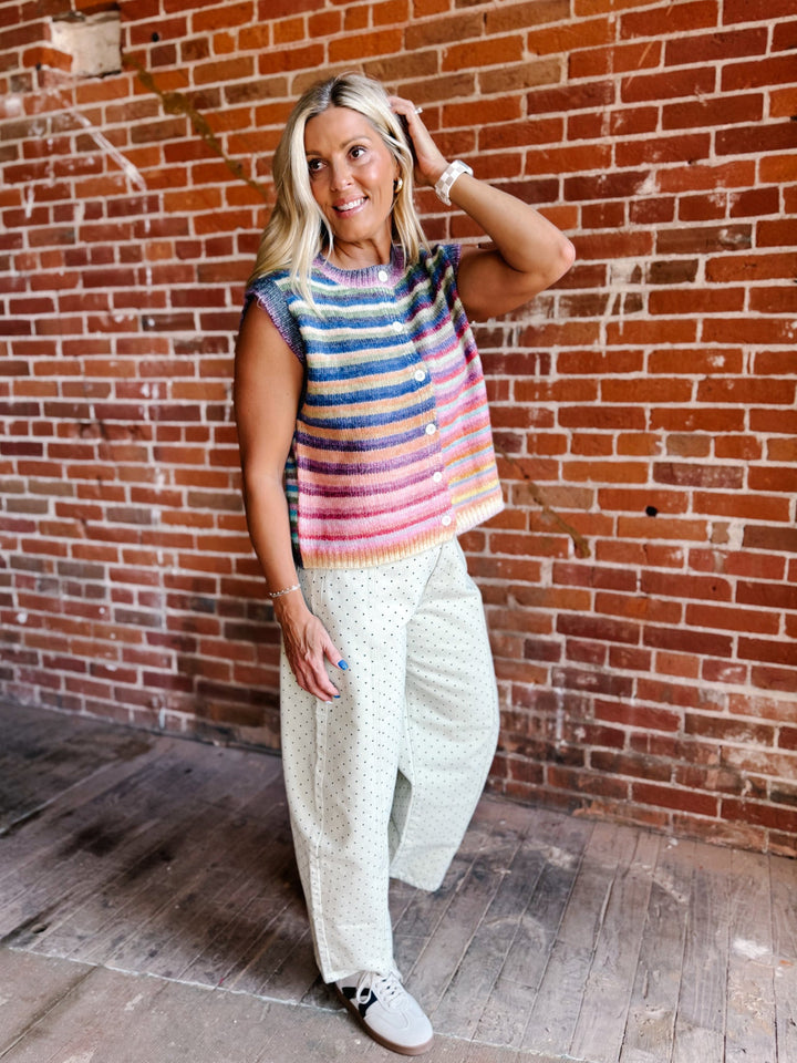 A woman posing in front of a red brick wall, wearing a colorful striped sleeveless knit top, white wide-leg patterned pants, and white sneakers.