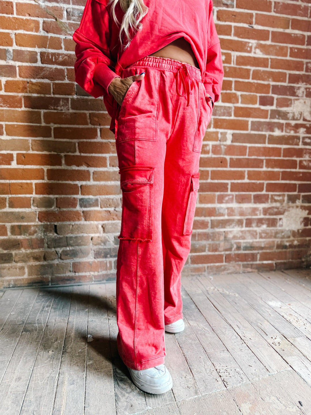A person wearing bright red wide-leg cargo pants and a matching red sweatshirt, standing against a brick wall and wooden floor.