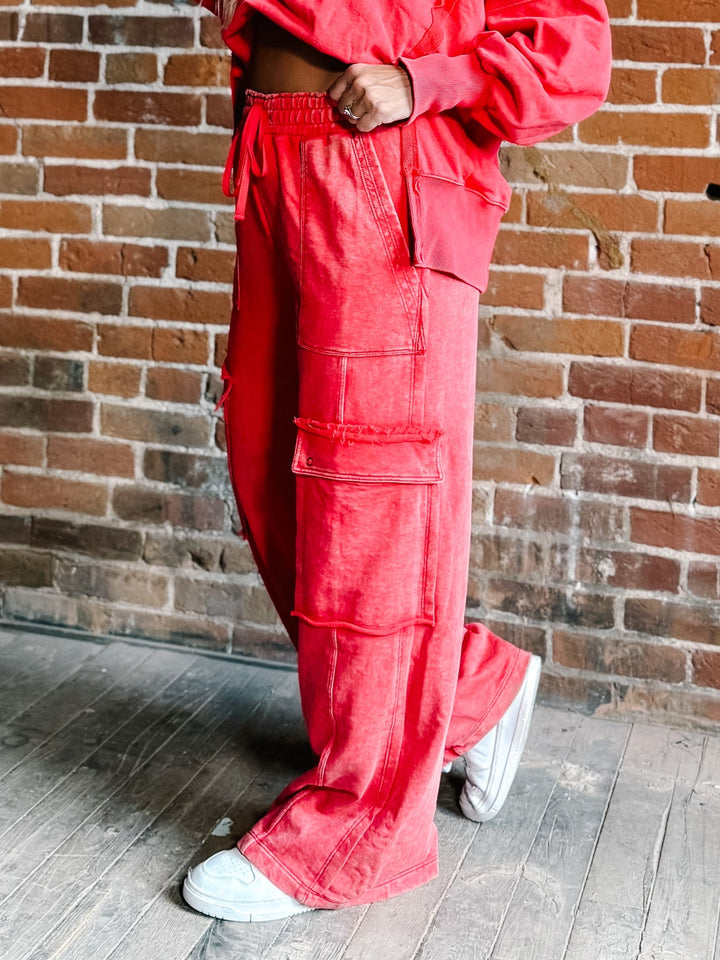 A person wearing bright coral-red wide-leg cargo pants with large flap pockets, paired with white sneakers, standing against a rustic brick wall.