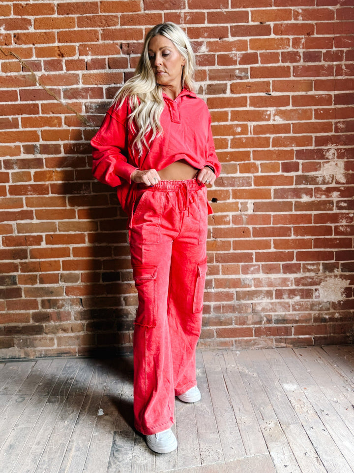 A woman with blonde hair stands against a red brick wall wearing a matching bright coral-red cropped long-sleeve top and wide-leg cargo pants, paired with white sneakers.