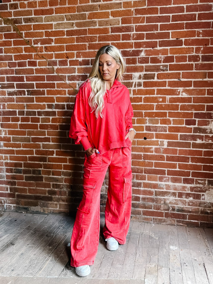 A woman with blonde hair stands against a red brick wall wearing a matching bright coral-red two-piece lounge set consisting of a cropped long-sleeve top and wide-leg pants, paired with white sneakers.