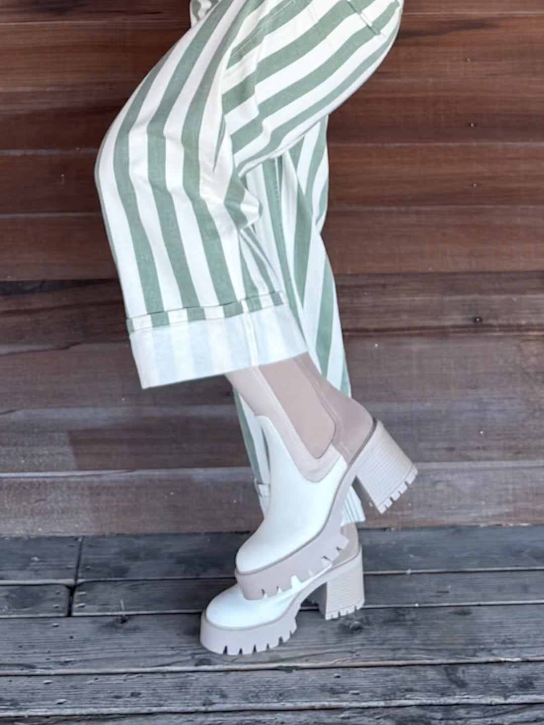 White platform Chelsea boots with chunky lug soles and elastic side panels, worn with green and white striped trousers against a rustic wooden plank background.