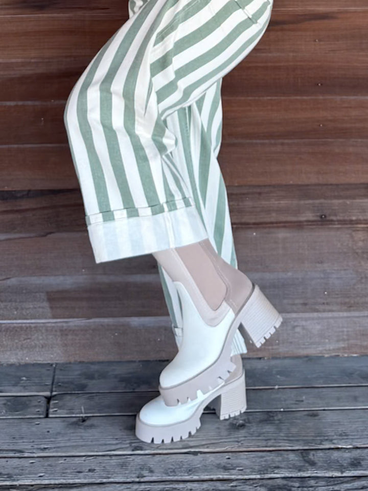 White platform Chelsea boots with chunky lug soles and elastic side panels, worn with green and white striped trousers against a rustic wooden plank background.