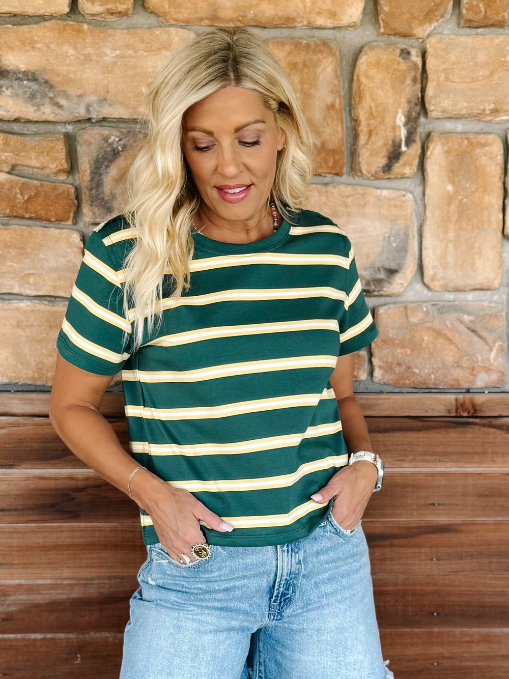 A woman wearing a green and yellow striped short sleeve top with a relaxed fit, standing against a stone wall.