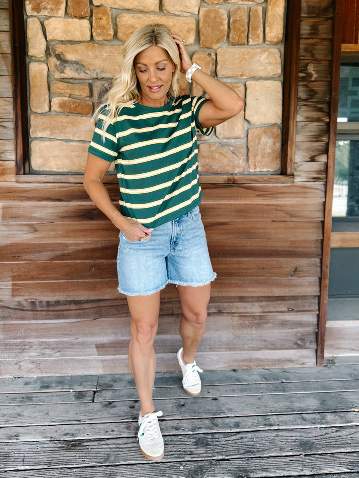 A woman wearing a green and yellow striped short sleeve top with a relaxed fit, standing against a stone wall.