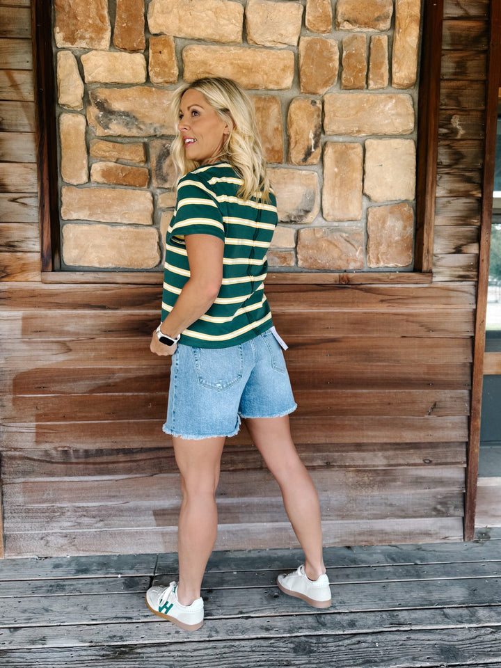 A woman wearing a green and yellow striped short sleeve top with a relaxed fit, standing against a stone wall.