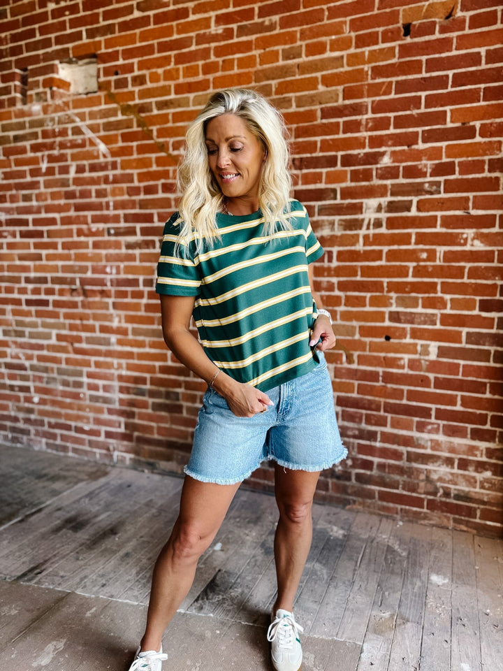 A woman wearing a green and yellow striped short sleeve top with a relaxed fit, standing against a stone wall.