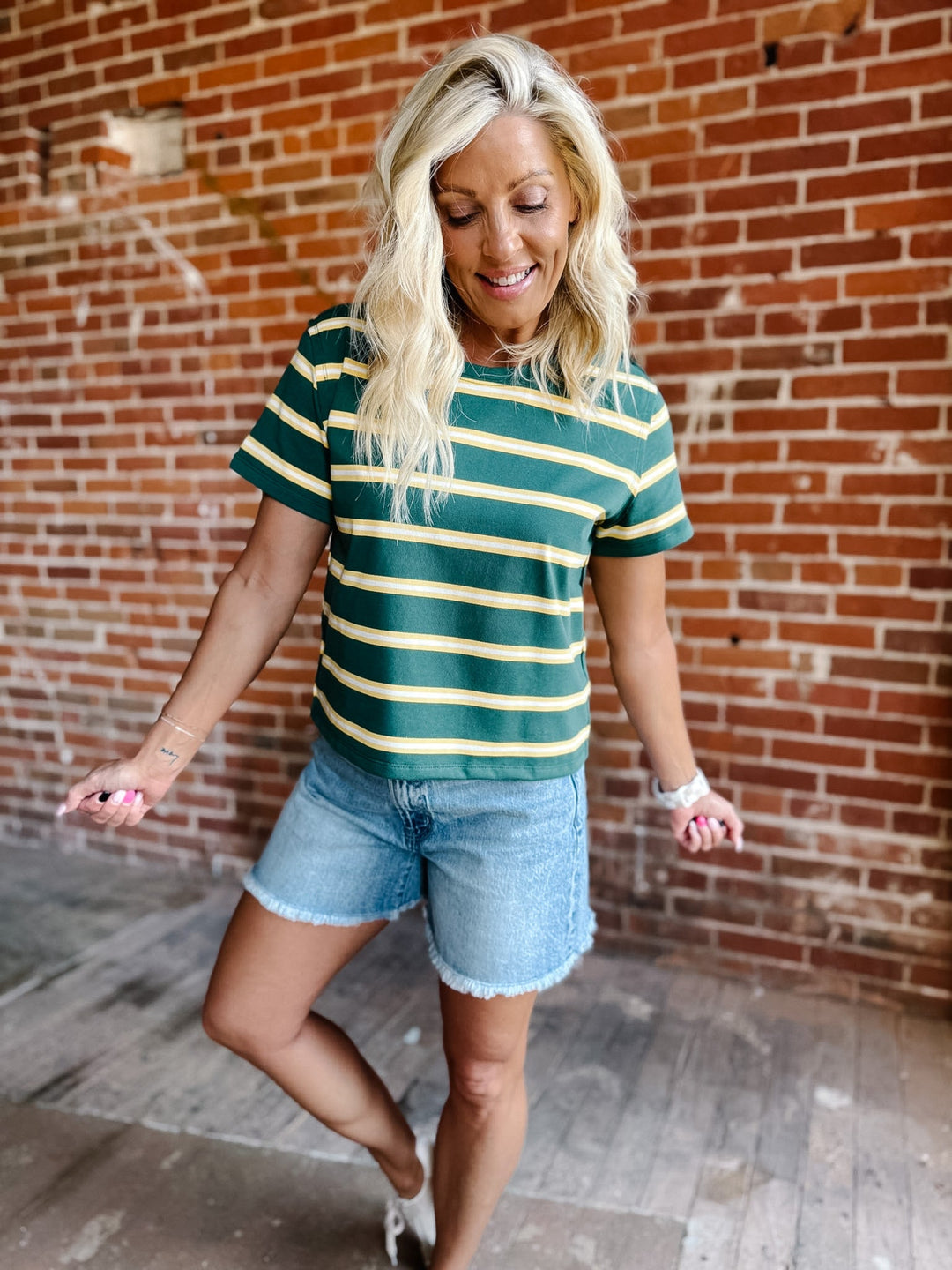 A woman wearing a green and yellow striped short sleeve top with a relaxed fit, standing against a stone wall.