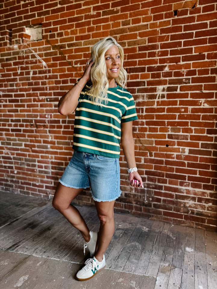 A woman wearing a green and yellow striped short sleeve top with a relaxed fit, standing against a stone wall.