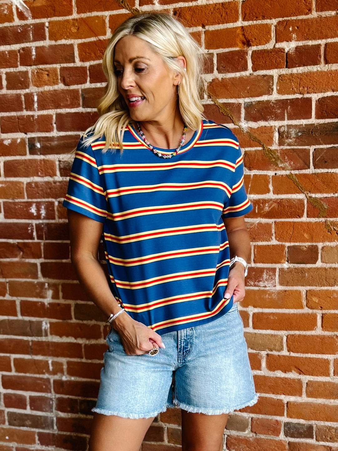 A woman wearing a blue, red and yellow striped short sleeve top with a relaxed fit, standing against a stone wall.