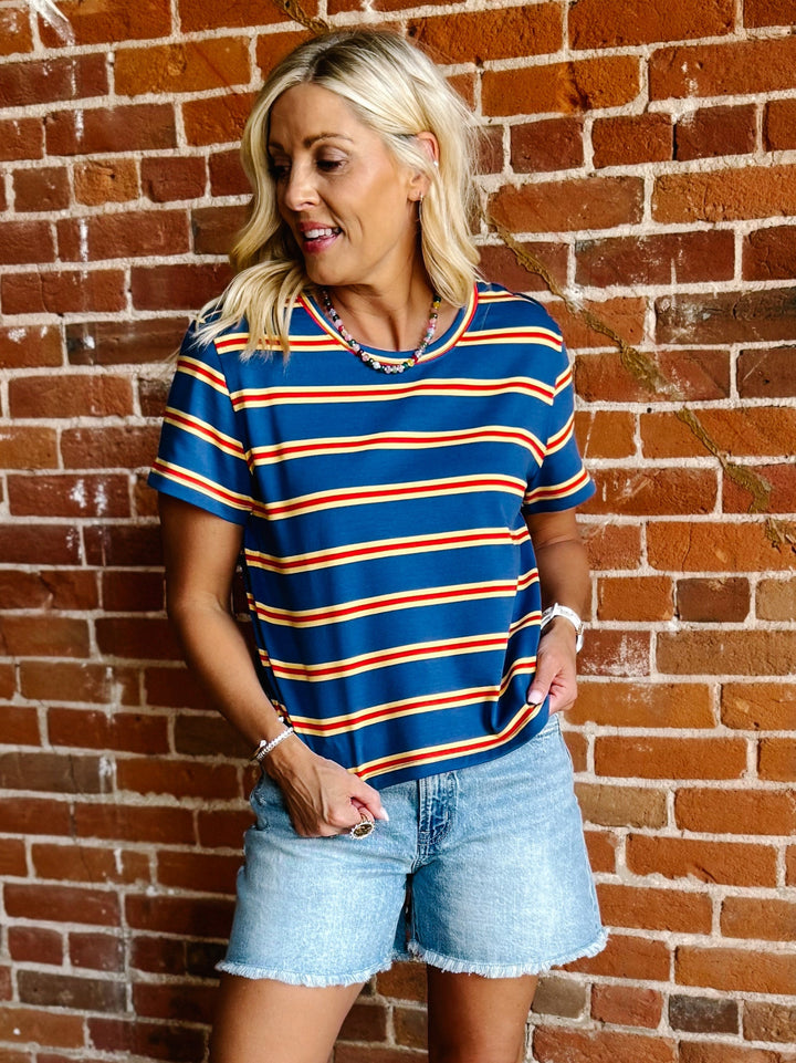 A woman wearing a blue, red and yellow striped short sleeve top with a relaxed fit, standing against a stone wall.