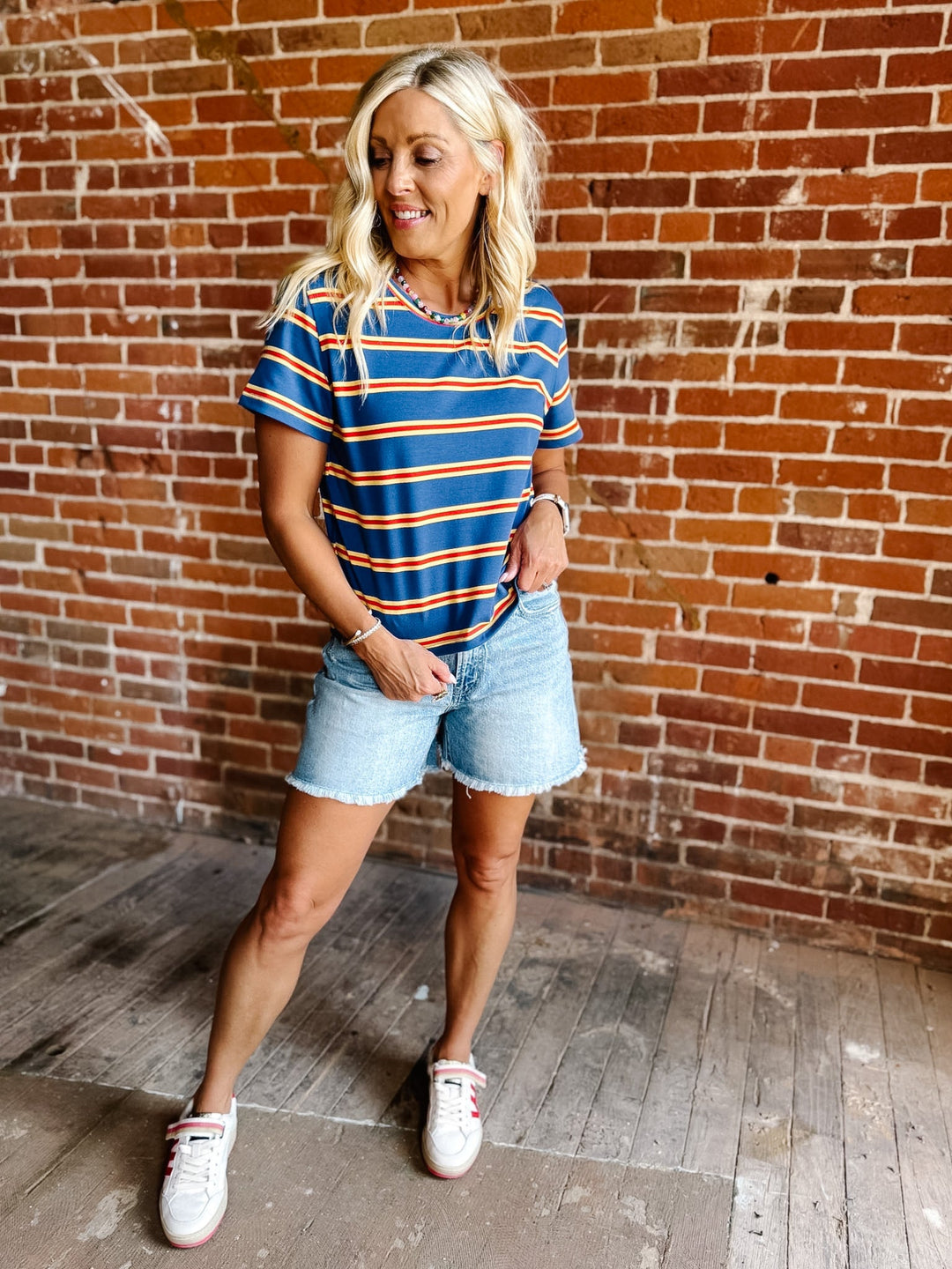 A woman wearing a blue, red and yellow striped short sleeve top with a relaxed fit, standing against a stone wall.