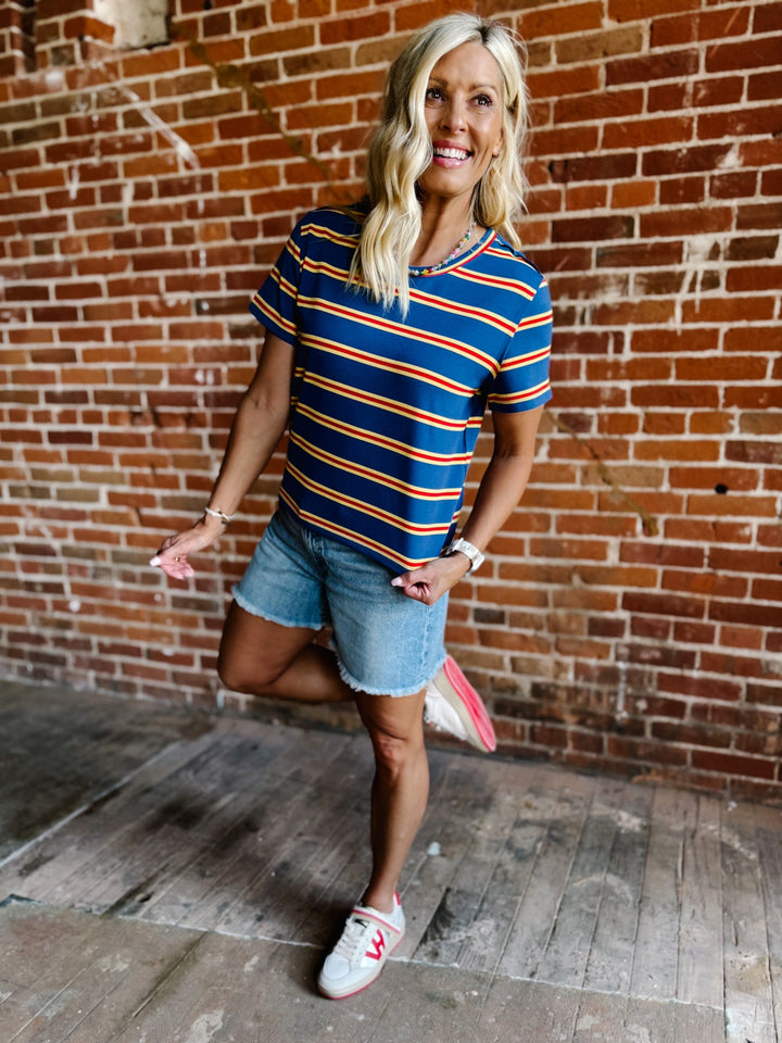 A woman wearing a blue, red and yellow striped short sleeve top with a relaxed fit, standing against a stone wall.