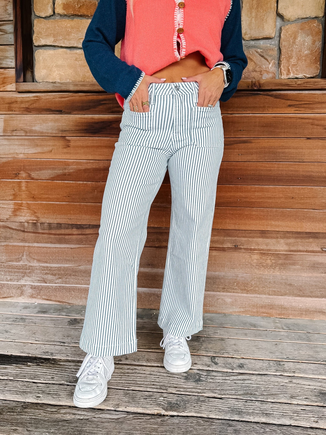 A person wearing blue and white vertically striped wide-leg pants, a coral sleeveless top, and a navy long-sleeve shirt, standing on a rustic wooden porch.
