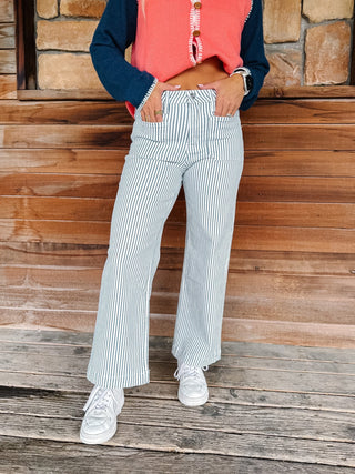 A person wearing blue and white vertically striped wide-leg pants, a coral sleeveless top, and a navy long-sleeve shirt, standing on a rustic wooden porch.