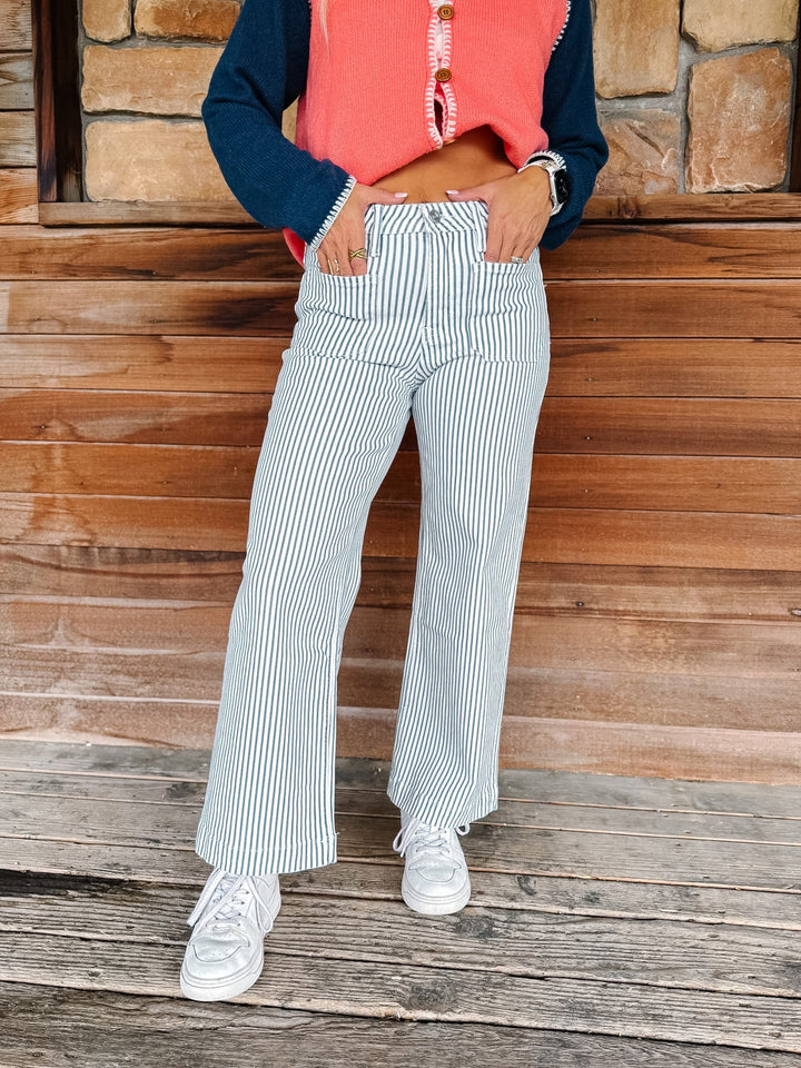 A person wearing blue and white vertically striped wide-leg pants, a coral sleeveless top, and a navy long-sleeve shirt, standing on a rustic wooden porch.