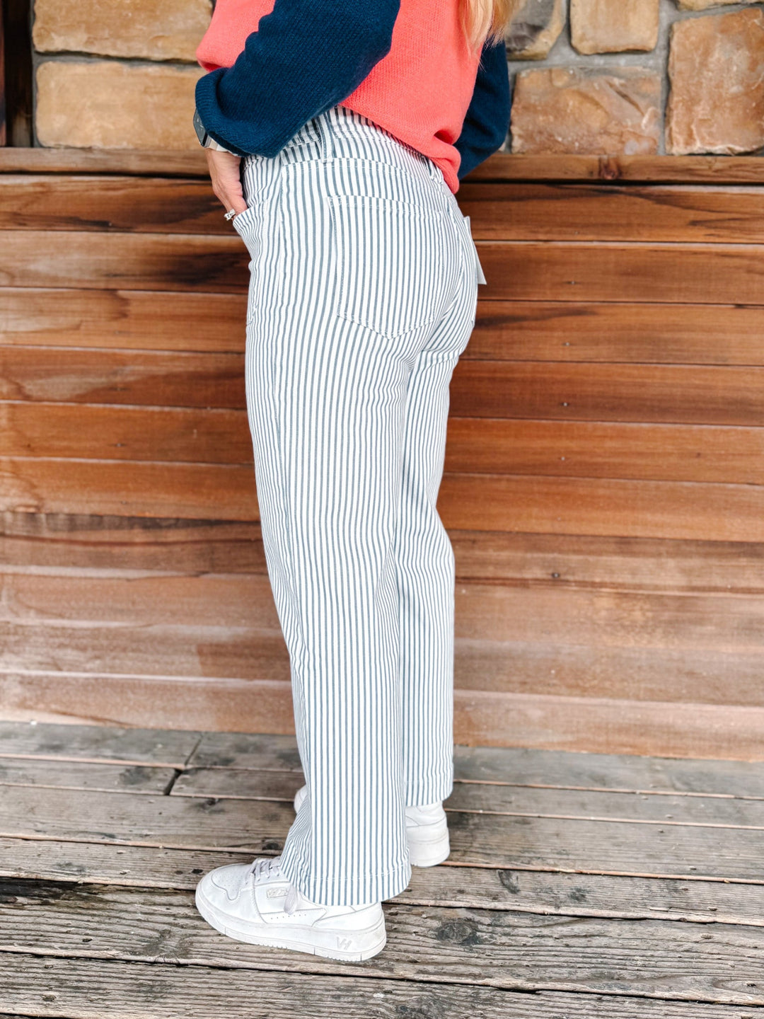 A person wearing light blue and white vertically striped wide-leg pants, a coral and navy cardigan, and white sneakers, standing against a rustic wooden wall and deck.
