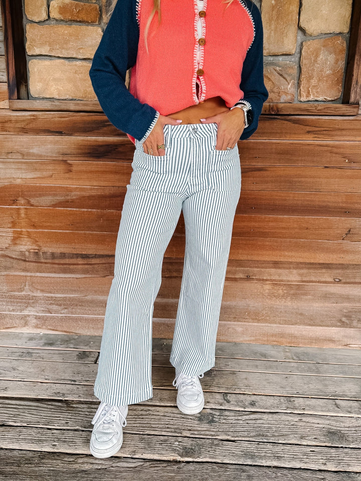 A person wearing striped wide-leg jeans, a coral and navy cardigan, and white sneakers, standing on a wooden deck in front of a stone wall.