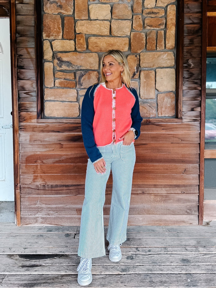 A woman stands on a wooden porch in front of a stone and wood wall, wearing a coral and navy cardigan, light blue and white vertically striped flared pants, and white sneakers.
