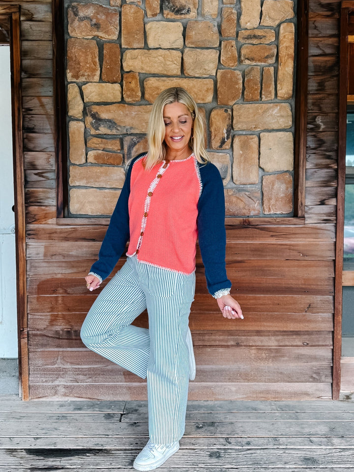 A woman posing on a rustic wooden porch in front of a stone wall, wearing a coral and navy raglan cardigan with striped pants.