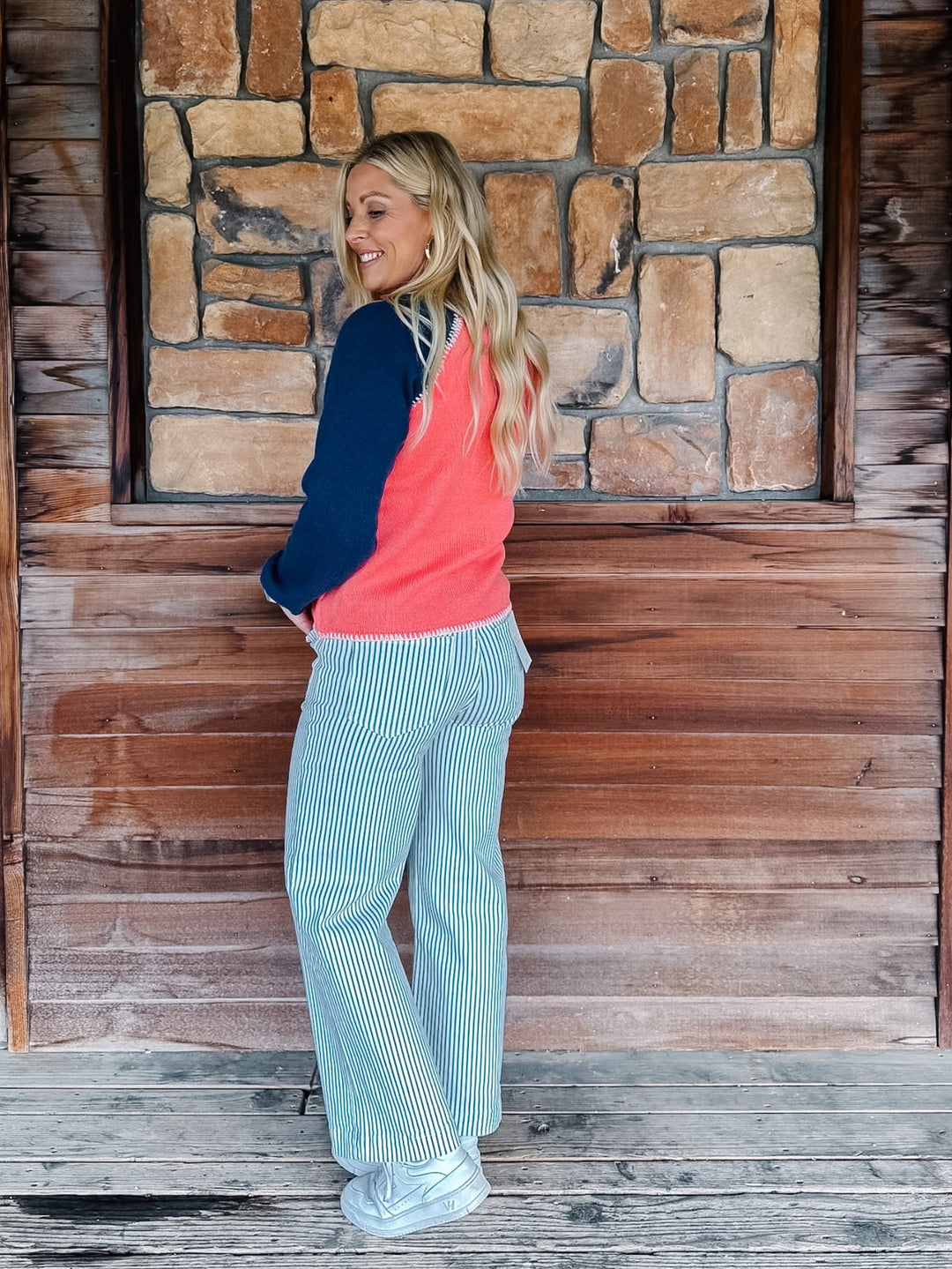 A woman stands on a wooden porch in front of a stone wall, wearing a coral and navy color-blocked cardigan and white vertically striped wide-leg pants.