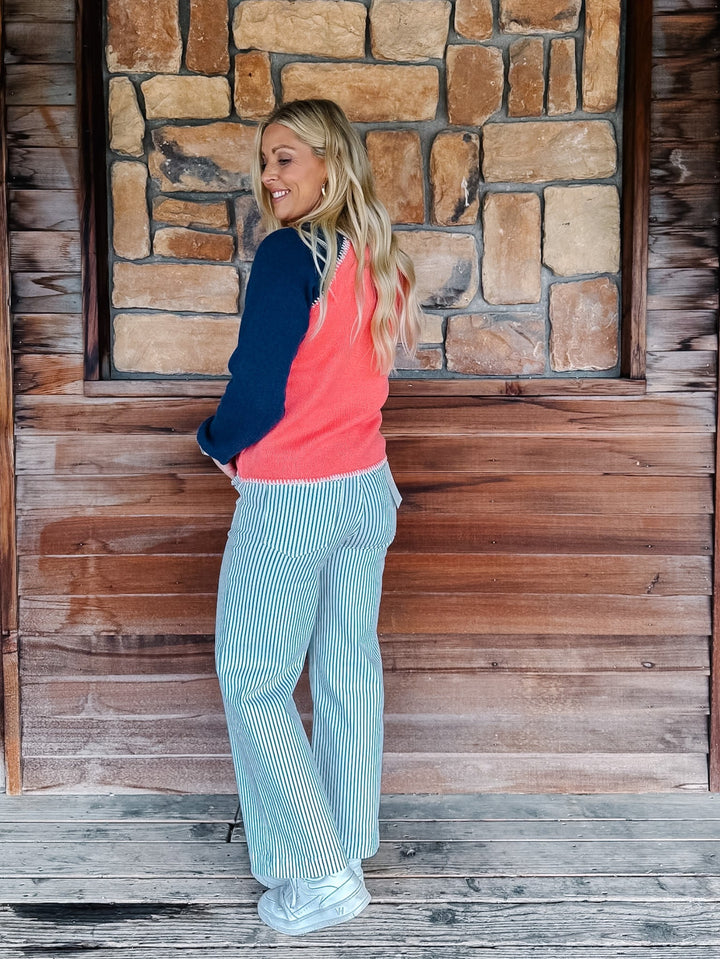 A woman stands on a wooden porch in front of a stone wall, wearing a coral and navy color-blocked cardigan and white vertically striped wide-leg pants.