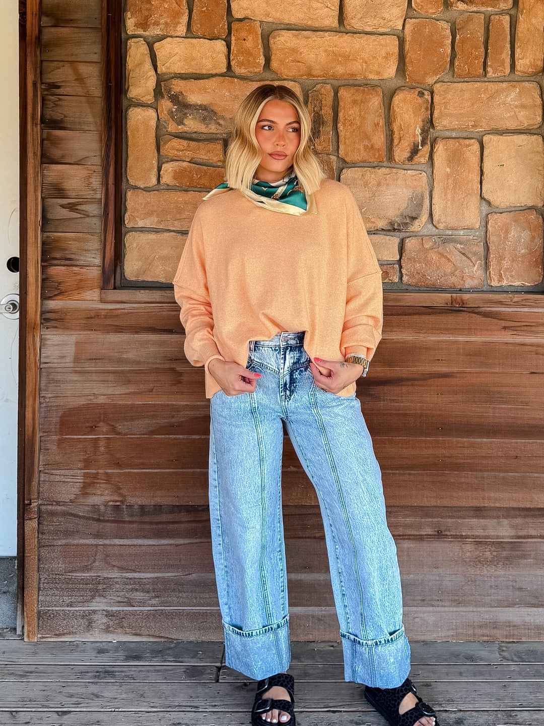 A woman stands on a wooden porch wearing a peach-colored knit sweater, light-wash wide-leg jeans, and black sandals, with a green and white patterned scarf tied around her neck.
