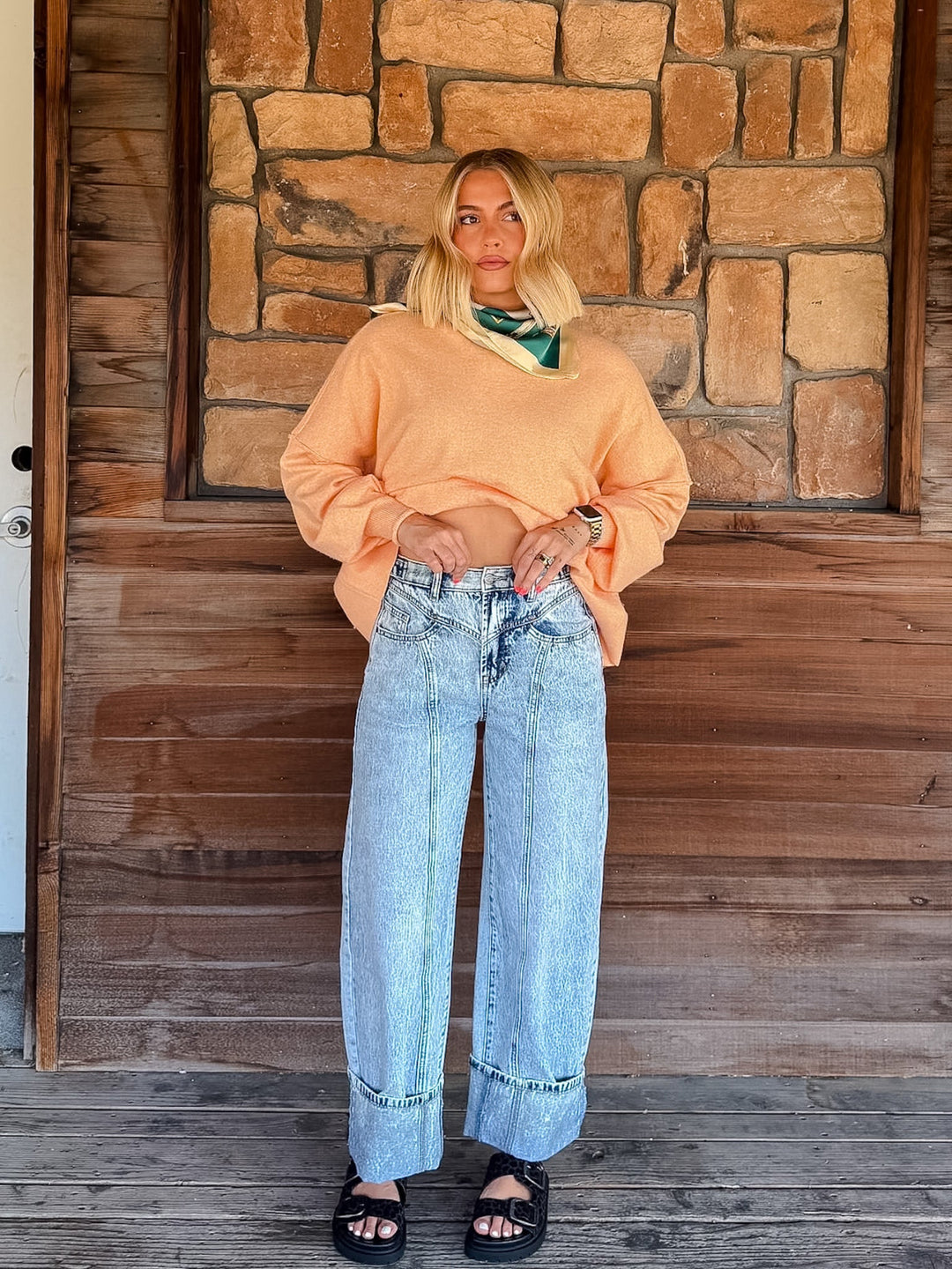 A woman stands on a wooden porch in front of a stone wall, wearing a light peach cropped knit sweater, light-wash wide-leg denim jeans, and black sandals.