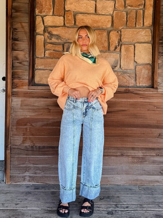 A woman stands on a wooden porch in front of a stone wall, wearing a light peach cropped knit sweater, light-wash wide-leg denim jeans, and black sandals.