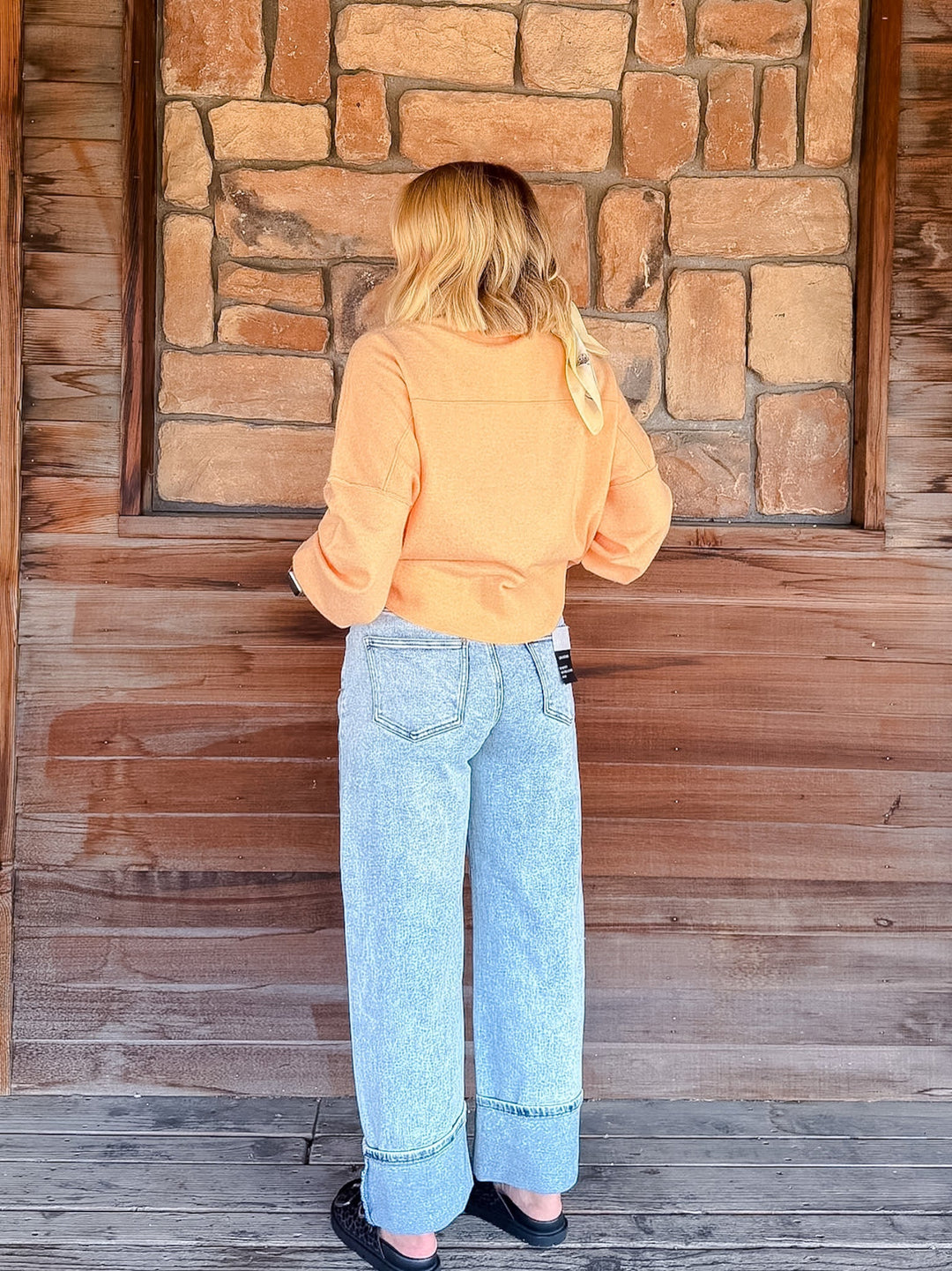 A woman viewed from behind wearing a light orange cropped long-sleeve top and light blue wide-leg jeans, standing in front of a rustic stone and wood wall.