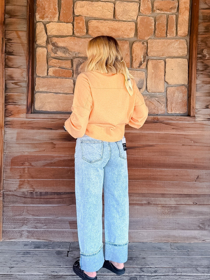 A woman viewed from behind wearing a light orange cropped long-sleeve top and light blue wide-leg jeans, standing in front of a rustic stone and wood wall.
