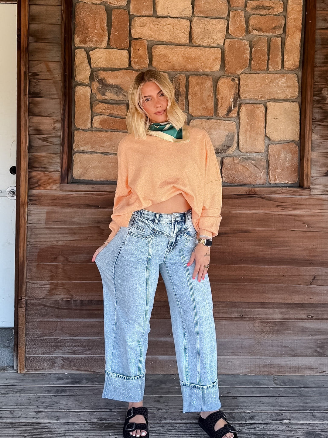 A woman stands on a wooden porch in front of a stone wall, wearing a peach cropped long-sleeve top, light-wash wide-leg jeans with rolled cuffs, and black sandals.
