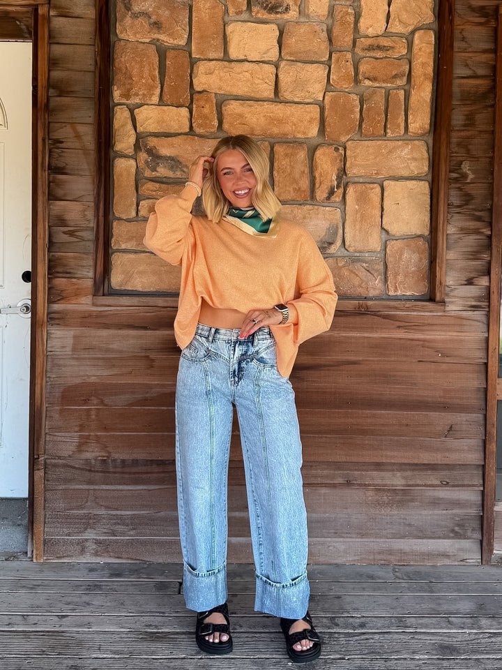 A woman stands on a wooden porch in front of a stone wall, wearing a cropped orange long-sleeve top, light blue wide-leg jeans, and black slide sandals.