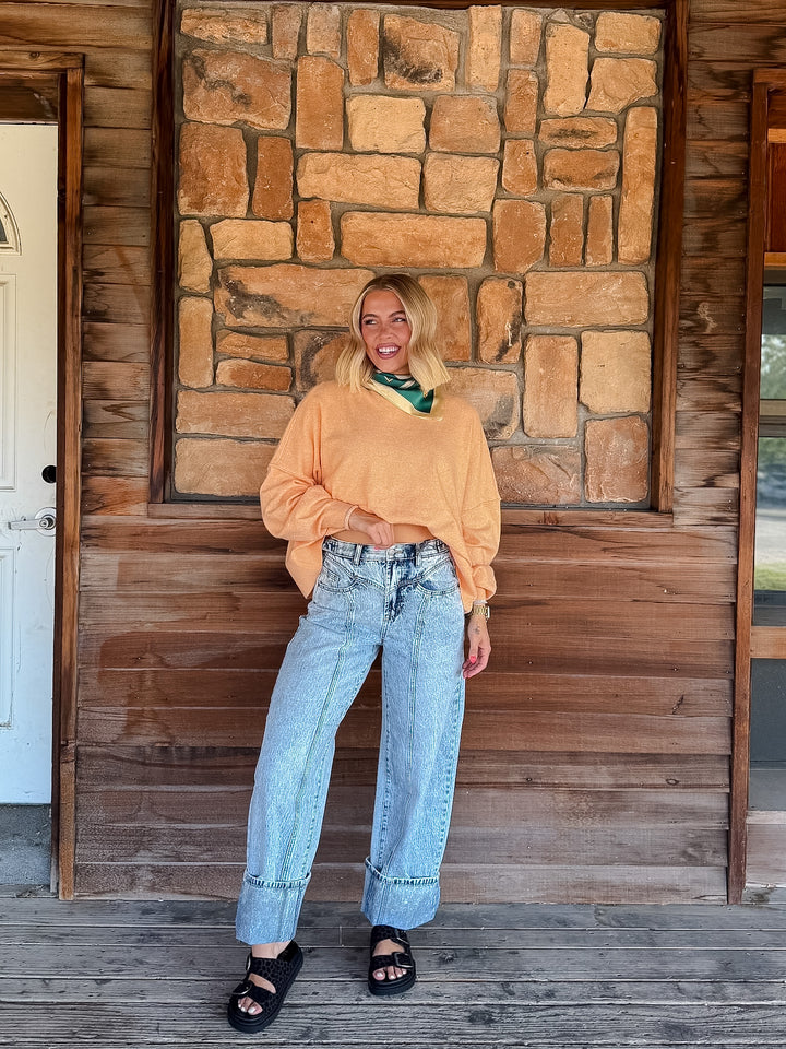 A woman stands on a wooden porch in front of a stone wall, wearing a light peach-orange long-sleeve sweatshirt, light-wash wide-leg jeans, and black slide sandals.