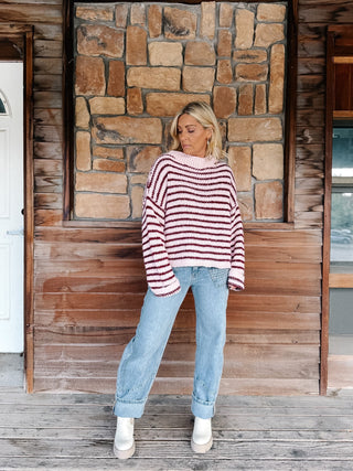 A woman wearing a chunky knit sweater with horizontal burgundy and pink stripes, paired with light blue jeans, standing in front of a rustic stone wall.