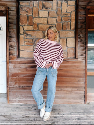 A woman wearing a chunky knit sweater with horizontal burgundy and pink stripes, paired with light blue jeans, standing in front of a rustic stone wall.