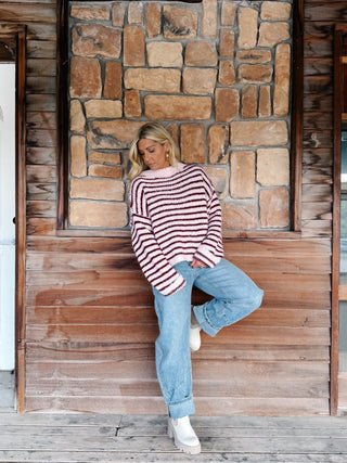 A woman wearing a chunky knit sweater with horizontal burgundy and pink stripes, paired with light blue jeans, standing in front of a rustic stone wall.