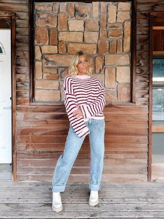 A woman wearing a chunky knit sweater with horizontal burgundy and pink stripes, paired with light blue jeans, standing in front of a rustic stone wall.