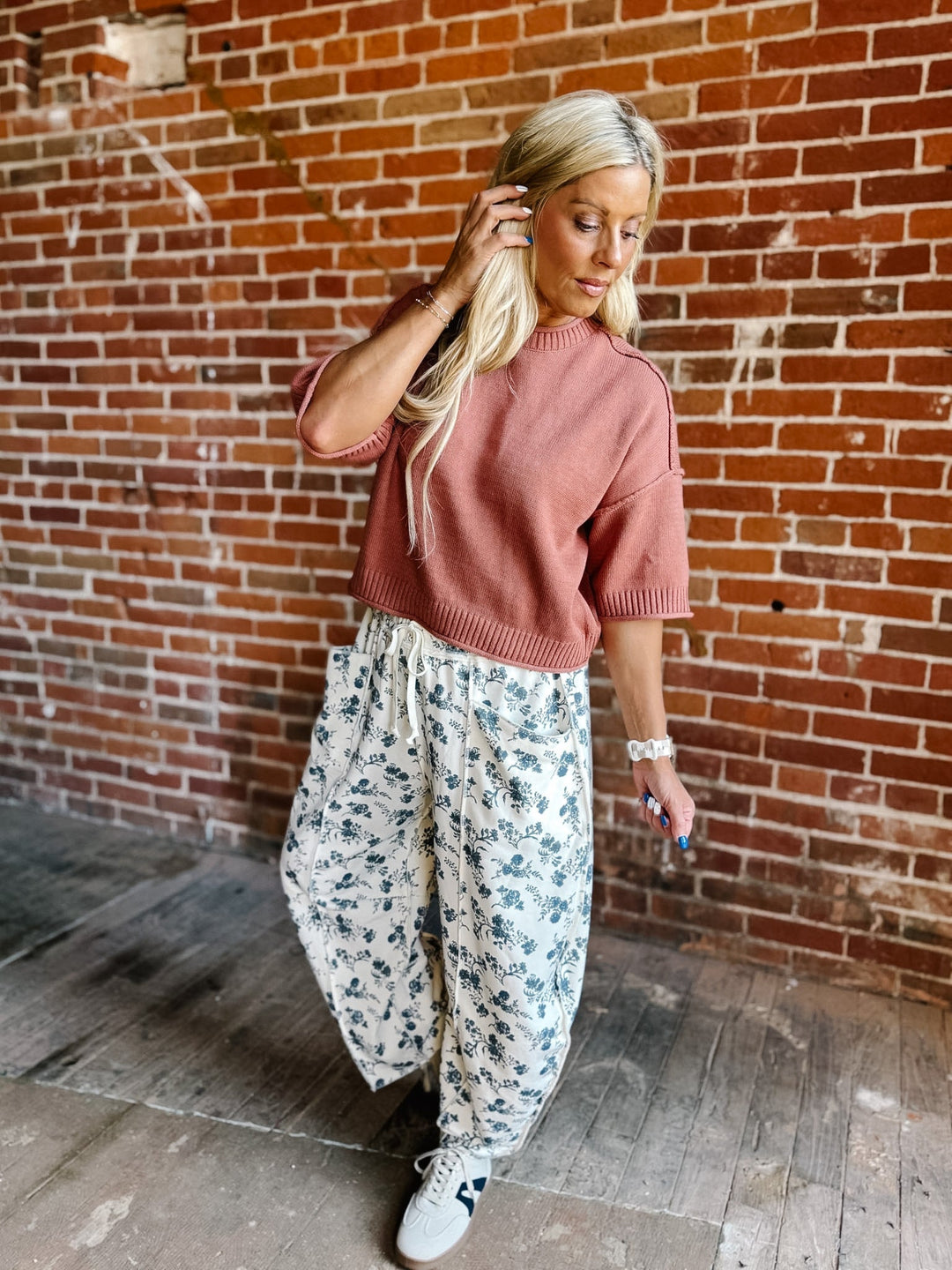 A woman stands in front of a red brick wall wearing a dusty rose short-sleeve knit sweater, white drawstring wide-leg pants with a blue floral print, and white sneakers.