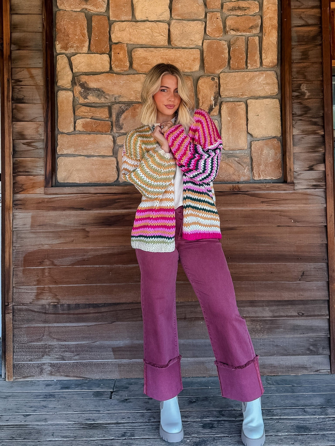 A woman stands on a rustic wooden porch in front of a stone wall, wearing a colorful striped knit cardigan and mauve wide-leg pants with white boots.
