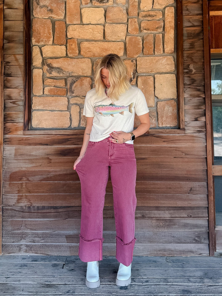 A woman stands on a wooden porch in front of a stone wall, wearing a cream graphic t-shirt, dusty rose wide-leg cropped jeans, and white platform sneakers.