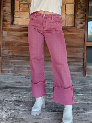 A person wearing dusty rose pink wide-leg cropped jeans with rolled cuffs, paired with white platform boots, standing on a weathered wooden deck in front of a rustic stone and wood wall.